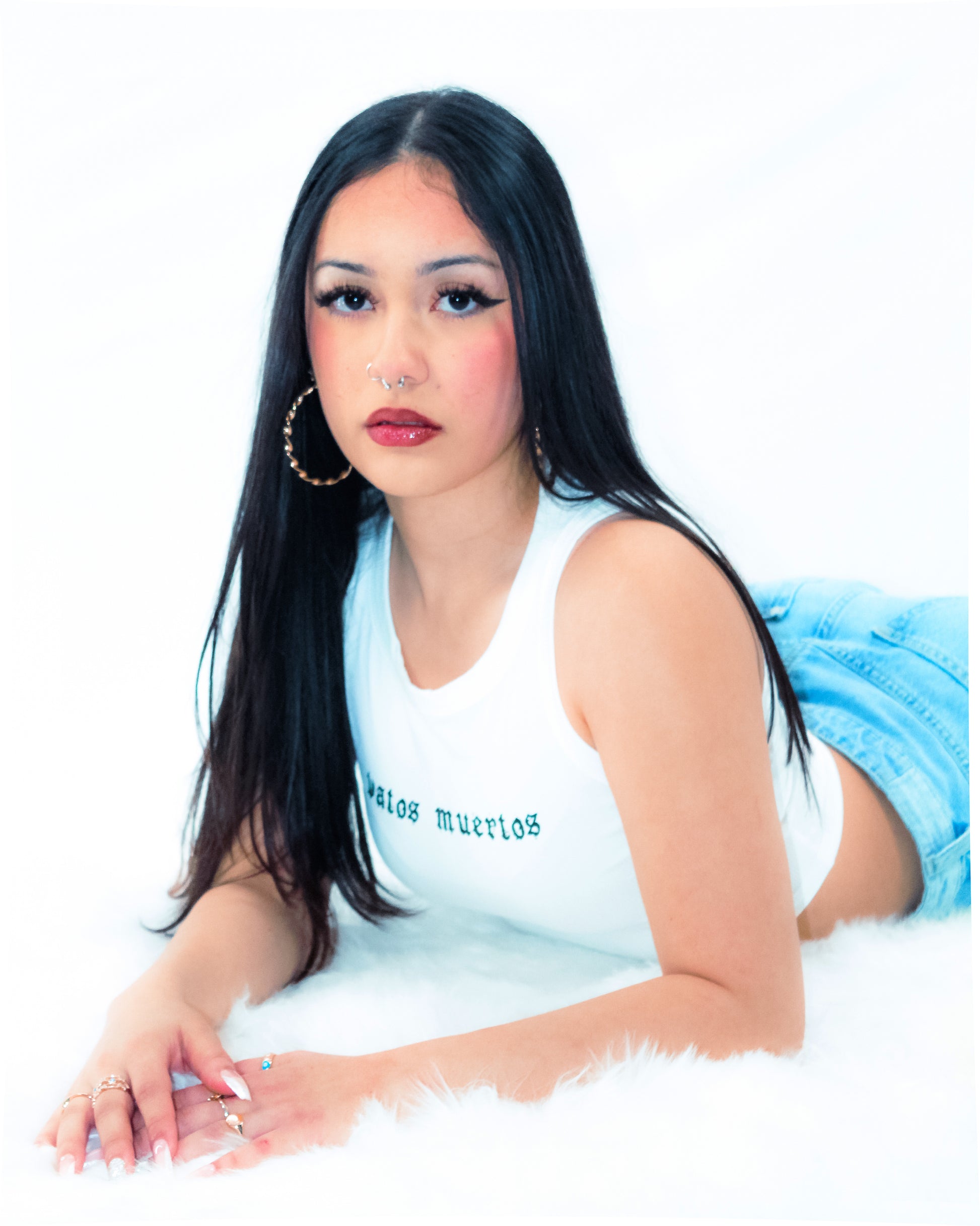 A woman with long black hair lies on a white faux-fur surface against a white studio background, wearing a white sleeveless top with green text and light blue denim shorts. She is also wearing large gold hoop earrings and rings.