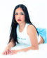 A woman with long black hair lies on a white faux-fur surface against a white studio background, wearing a white sleeveless top with green text and light blue denim shorts. She is also wearing large gold hoop earrings and rings.